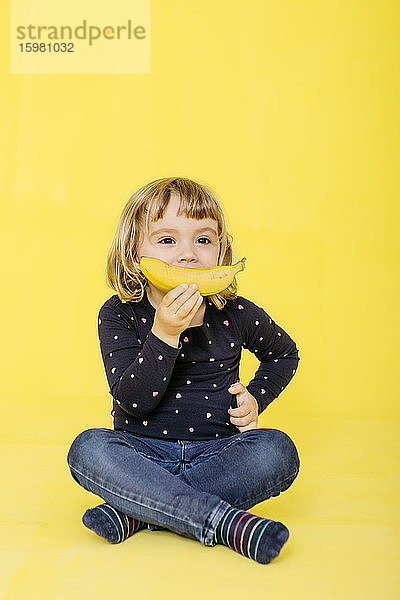 Vorschulmädchen in voller Länge  das eine Banane hält  während es im Schneidersitz sitzt und vor einem farbigen Hintergrund wegschaut