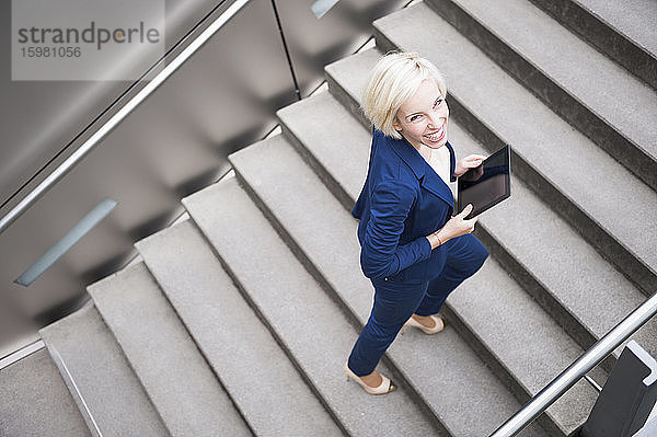 Porträt einer lächelnden blonden Geschäftsfrau beim Treppensteigen mit Tablet