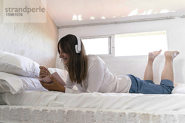 Lächelnde Frau auf dem Bett liegend  die mit Kopfhörern Musik hört und Notizen macht