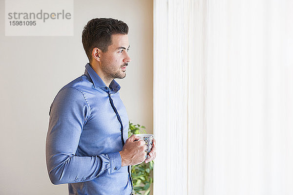 Nachdenklicher Mann  der eine Kaffeetasse hält  während er zu Hause am Fenster steht