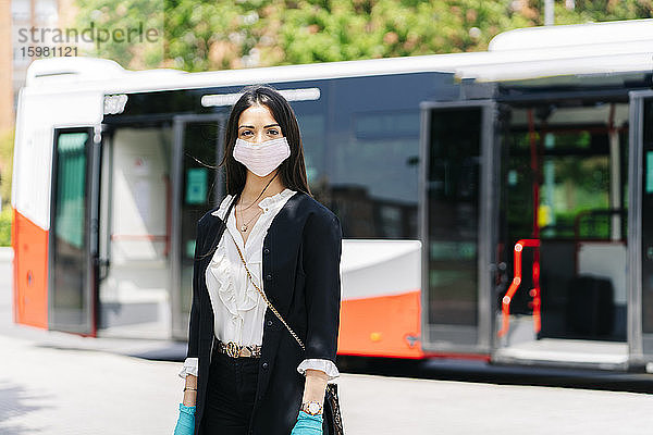 Porträt einer jungen Frau mit Schutzmaske und Handschuhen  die an einer Bushaltestelle wartet  Spanien