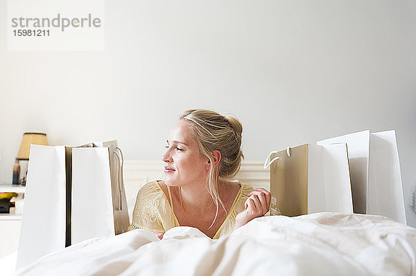 Porträt einer lächelnden blonden Frau auf dem Bett zwischen Einkaufstüten