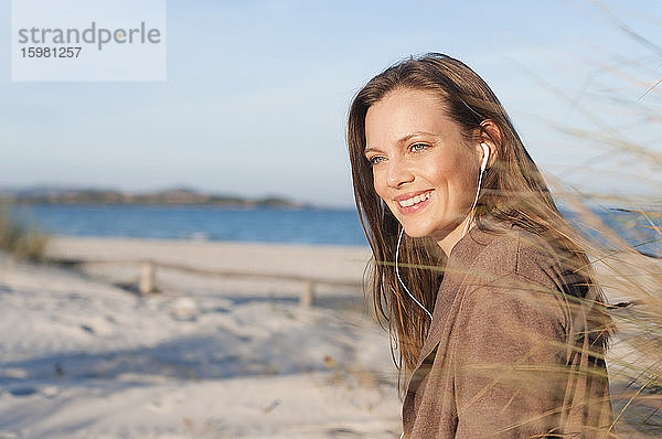 Porträt einer lächelnden Frau am Strand  die mit Kopfhörern Musik hört  Sardinien  Italien