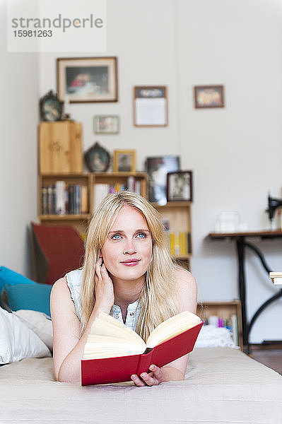 Porträt einer lächelnden blonden Frau  die mit einem Buch auf der Couch liegt und in die Ferne schaut