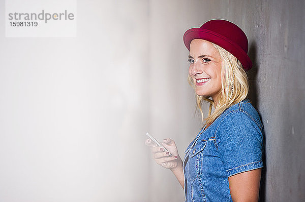Porträt einer glücklichen jungen Frau mit Smartphone  die an der Wand lehnt