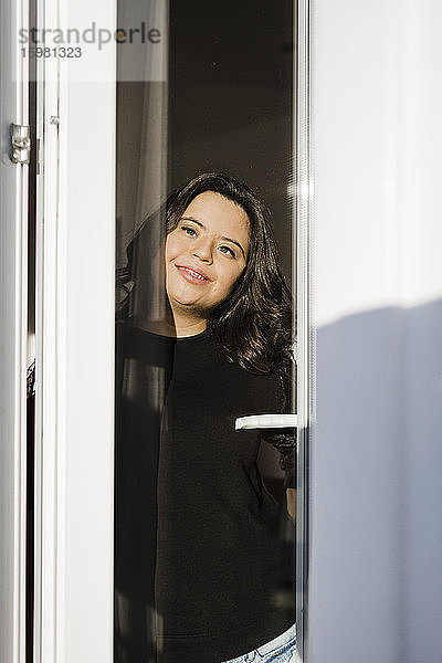 Nachdenkliche Frau mit Down-Syndrom lächelt  während sie durch das Fenster ihres Hauses schaut