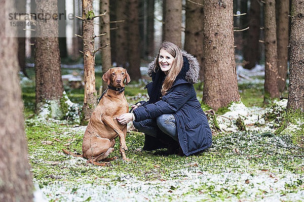 Lächelnde junge schöne Frau  die mit ihrem Hund vor einem Baum im Wald kauert