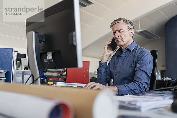 Gutaussehender männlicher Fachmann  der am Computerschreibtisch im Büro sitzt und mit einem Mobiltelefon spricht
