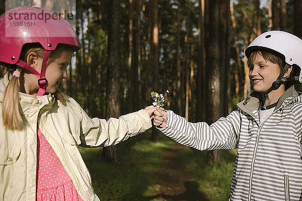 Mädchen und Junge mit Fahrradhelmen halten Blumen im Wald