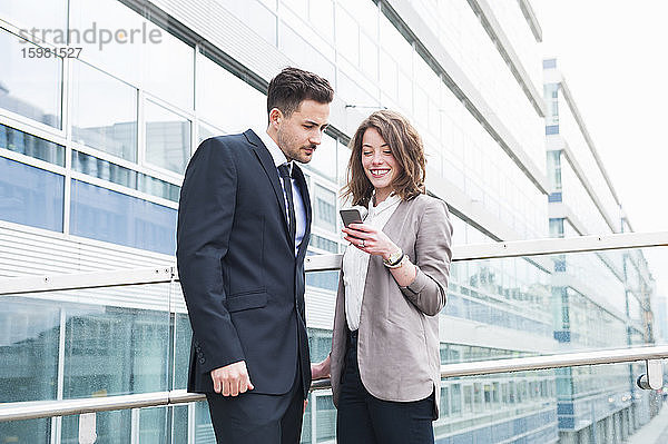 Glückliches Geschäftspaar benutzt ein Smartphone  während es vor einem Bürogebäude steht