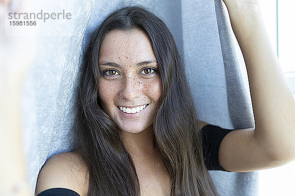 Nahaufnahme einer lächelnden jungen Frau mit Sommersprossen vor einem Vorhang zu Hause