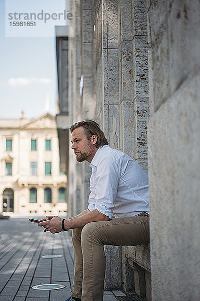 Geschäftsmann sitzt in der Nische eines Gebäudes und hält ein Smartphone