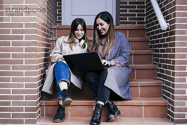 Porträt von zwei Frauen  die auf einer Treppe im Freien sitzen und auf einen Laptop schauen