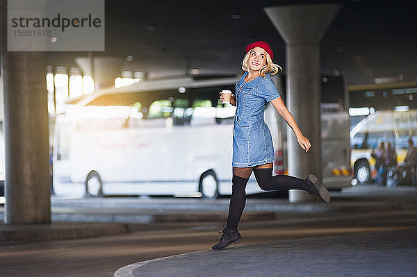Porträt einer glücklichen jungen Frau mit Kaffee zum Mitnehmen  die am Busbahnhof in die Luft springt