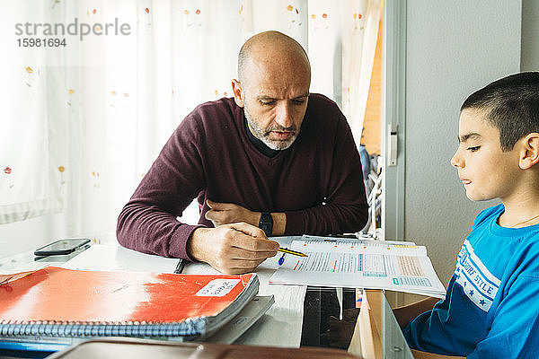 Selbstbewusster Vater unterrichtet seinen Sohn am Schreibtisch sitzend während des Homeschoolings
