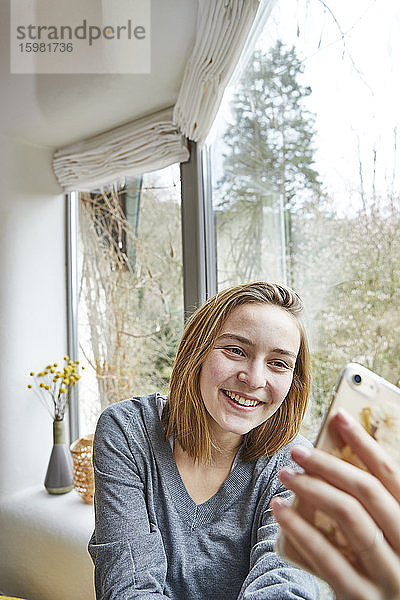Porträt einer lächelnden jungen Frau  die zu Hause ein Selfie mit ihrem Smartphone macht