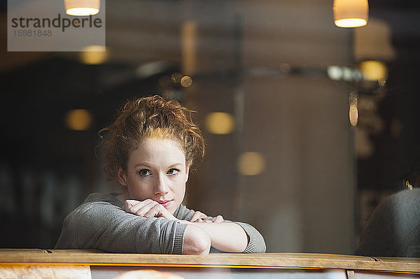 Nachdenkliche junge Frau  die sich auf einen Tisch stützt  gesehen durch ein Glasfenster in einem Café