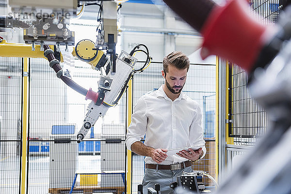 Junger männlicher Experte verwendet ein digitales Tablet  während er an einem Roboterarm in der Industrie steht