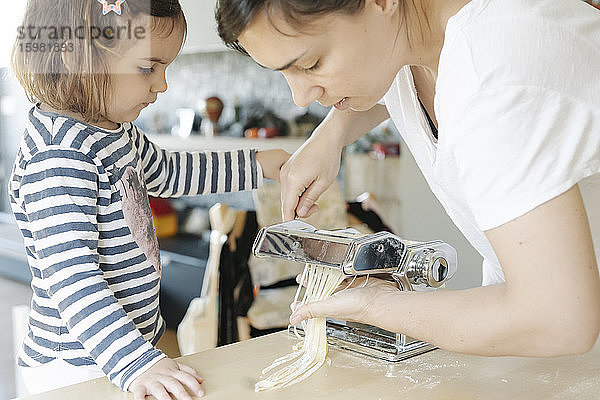 Nettes Mädchen schaut Mutter an  die Spaghetti mit Hilfe von Geräten in der Küche zubereitet
