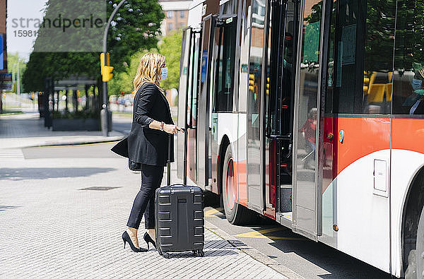 Geschäftsfrau mit Schutzmaske vor einem Bus mit Trolley-Tasche  Spanien