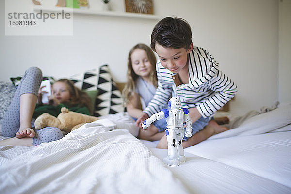 Aufgeregter kleiner Junge  der auf dem Bett hockt und mit einem Spielzeugroboter spielt  während sich seine Schwestern im Hintergrund entspannen