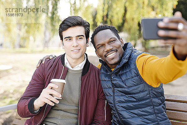 Zwei glückliche junge Männer sitzen auf einer Parkbank und machen ein Selfie