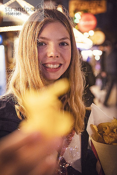 Porträt eines lächelnden blonden Mädchens  das beim nächtlichen Karneval in der Stadt Kartoffelchips isst. München  Deutschland