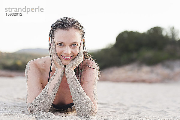 Porträt einer lächelnden Frau  die mit dem Kopf in den Händen am Strand liegt  Sardinien  Italien