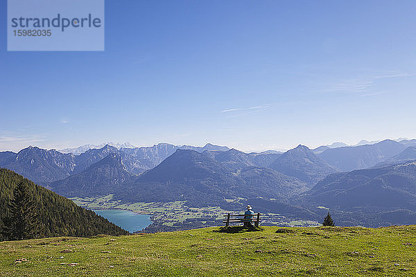 Rückansicht eines älteren männlichen Wanderers  der auf einer Bank sitzt und das Dachsteingebirge vor blauem Himmel betrachtet