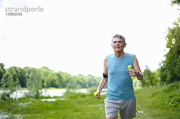 Aktiver älterer Mann joggt mit Hanteln am Flussufer gegen den klaren Himmel