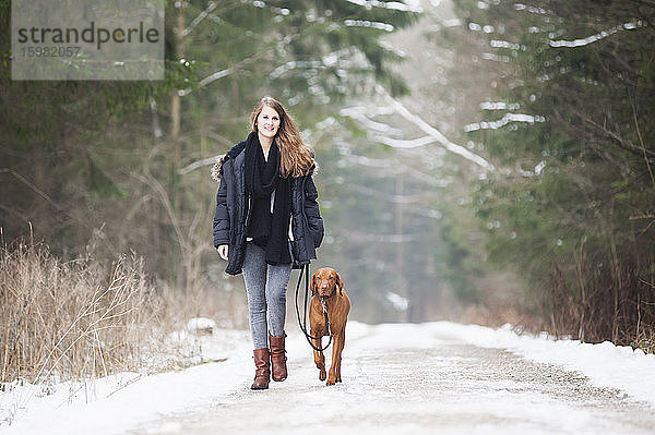 Schöne junge Frau geht mit Hund auf der Straße inmitten von Bäumen im Wald im Winter