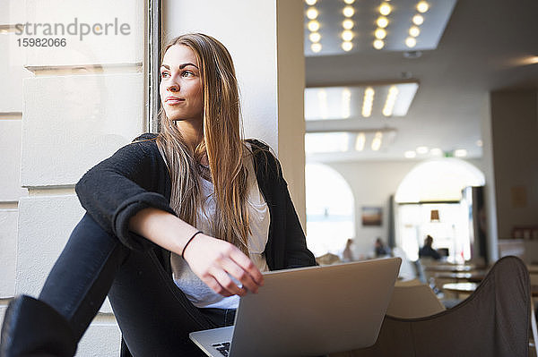 Nachdenkliche junge Frau sitzt mit Laptop auf der Fensterbank eines Cafés