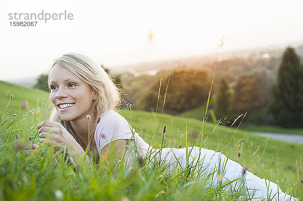 Lächelnde nachdenkliche junge Frau  die im Park bei Sonnenuntergang auf einer Wiese vor einem klaren Himmel liegt
