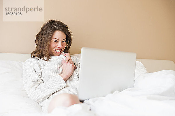 Glückliche junge Frau mit Laptop im Bett zu Hause