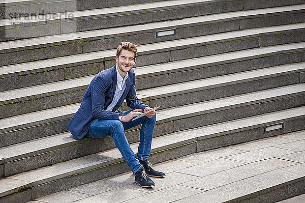 Glücklicher junger Geschäftsmann sitzt auf einer Treppe und benutzt ein Tablet