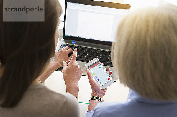 Rückenansicht einer älteren Frau und ihrer erwachsenen Tochter bei der Nutzung von Laptop und Smartphone