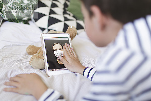 Auf dem Bett liegender Junge  der mit seiner Mutter  die eine Schutzmaske trägt  ein Videogespräch auf einem digitalen Tablet führt