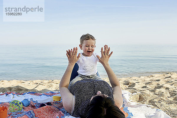 Mutter spielt mit ihrem Sohn ein Klatschspiel  während sie auf einer Decke am Strand liegt und das Meer sieht