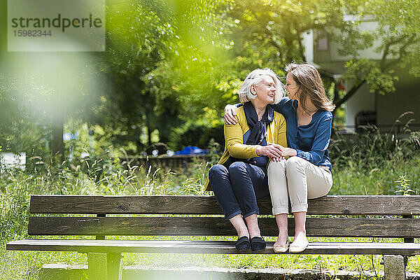 Senior woman and adult daughter sitting on backrest of park bench looking at each other