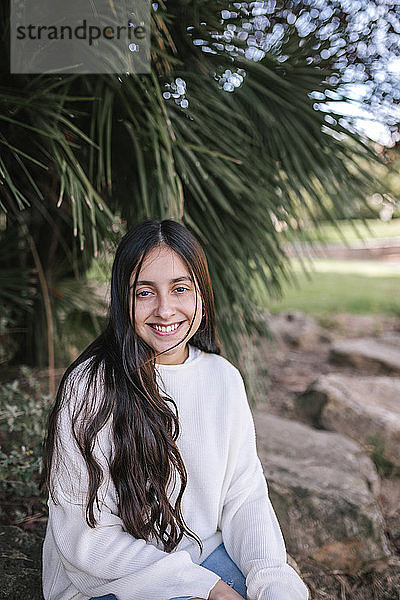 Porträt eines lächelnden Teenagers  der im Park vor einer Palme sitzt