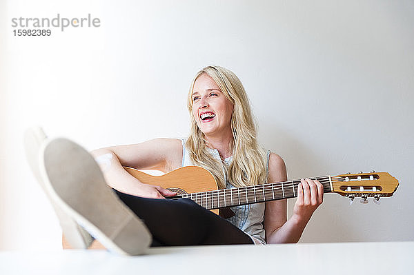 Porträt einer lachenden blonden Frau mit hochgelegten Füßen  die Gitarre spielt