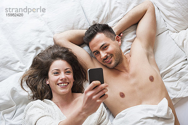 Fröhliche Frau  die ein Selfie mit einem Mann macht  während sie zu Hause im Bett liegt