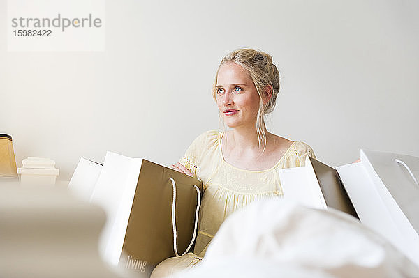 Porträt einer glücklichen blonden Frau  die zwischen Einkaufstüten auf dem Bett sitzt
