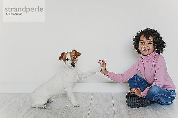 Porträt eines lächelnden kleinen Mädchens  das mit seinem Hund auf dem Boden sitzt und Pfötchen gibt