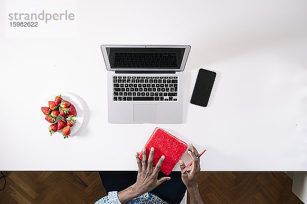 Junger Mann hält rotes Tagebuch neben Laptop und Smartphone mit Erdbeeren am Schreibtisch