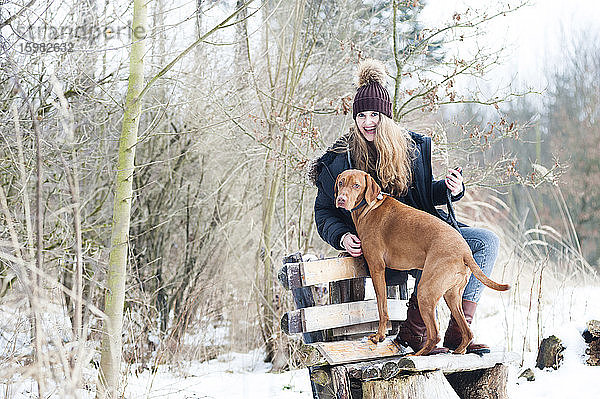 Fröhliche junge Frau mit Hund auf einer Bank sitzend vor kahlen Bäumen im Wald im Winter