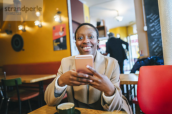 Glückliche junge Frau hält ihr Smartphone in der Hand  während sie in einem Café sitzt