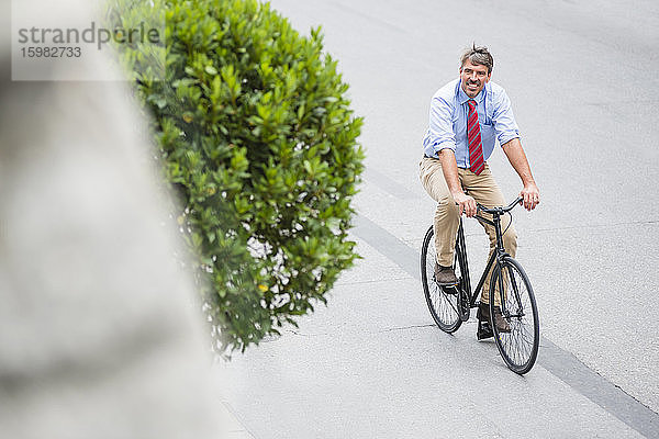 Lächelnder Geschäftsmann  der beim Fahrradfahren auf einer Straße in der Stadt nach oben schaut