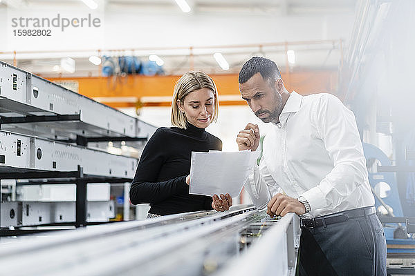 Geschäftsmann und Frau mit Papieren untersuchen Metallstangen in einer Fabrikhalle