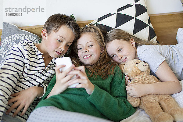 Gruppenbild von drei Kindern  die zusammen auf einem Bett liegen und auf ein Mobiltelefon schauen